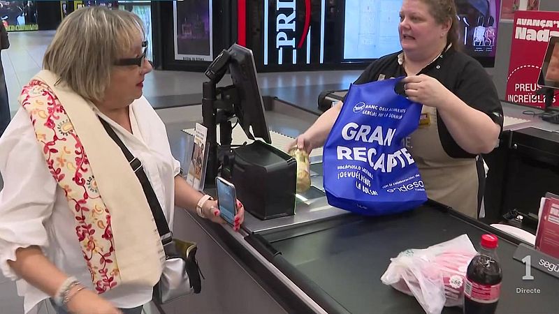 Clienta y cajera en el mostrador de un supermercado durante la campaña benéfica del Banc dels Aliments, con bolsas de productos y cartelería promocional de fondo.