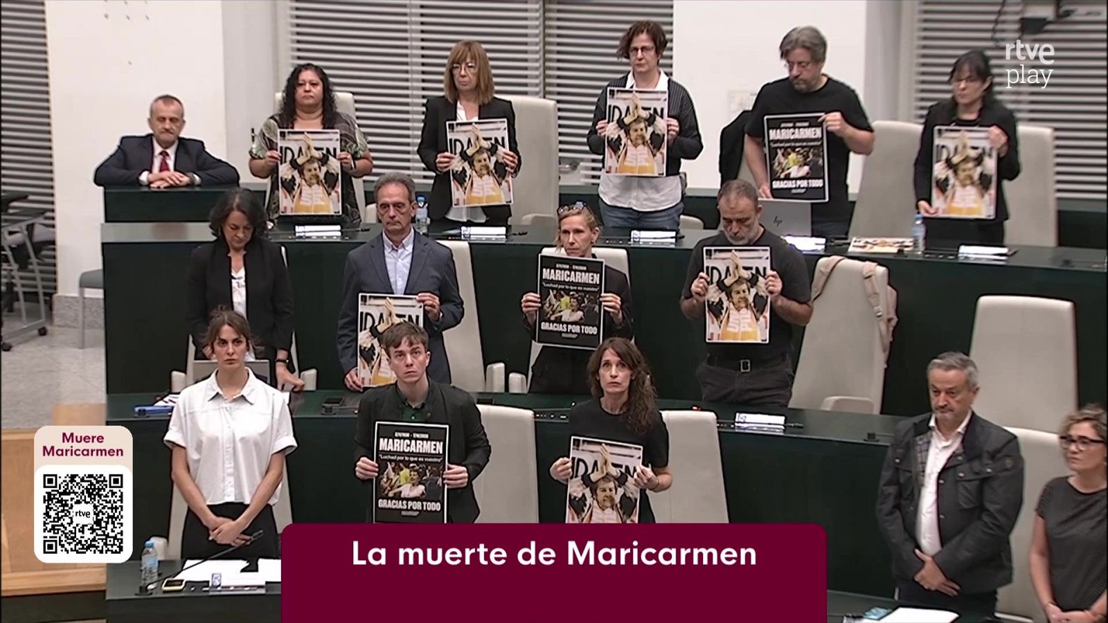 Representantes públicos guardan un minuto de silencio en un salón de plenos sosteniendo carteles de homenaje con la imagen de una artista fallecida.