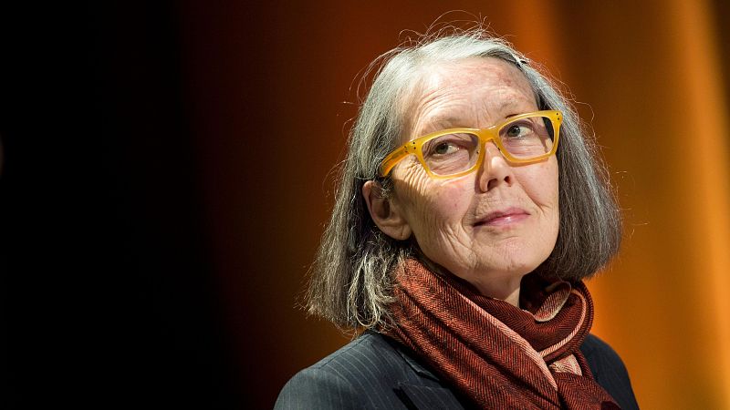 Retrato de la escritora Anne Carson con cabello canoso, gafas anaranjadas, chaqueta oscura de rayas y pañuelo granate.