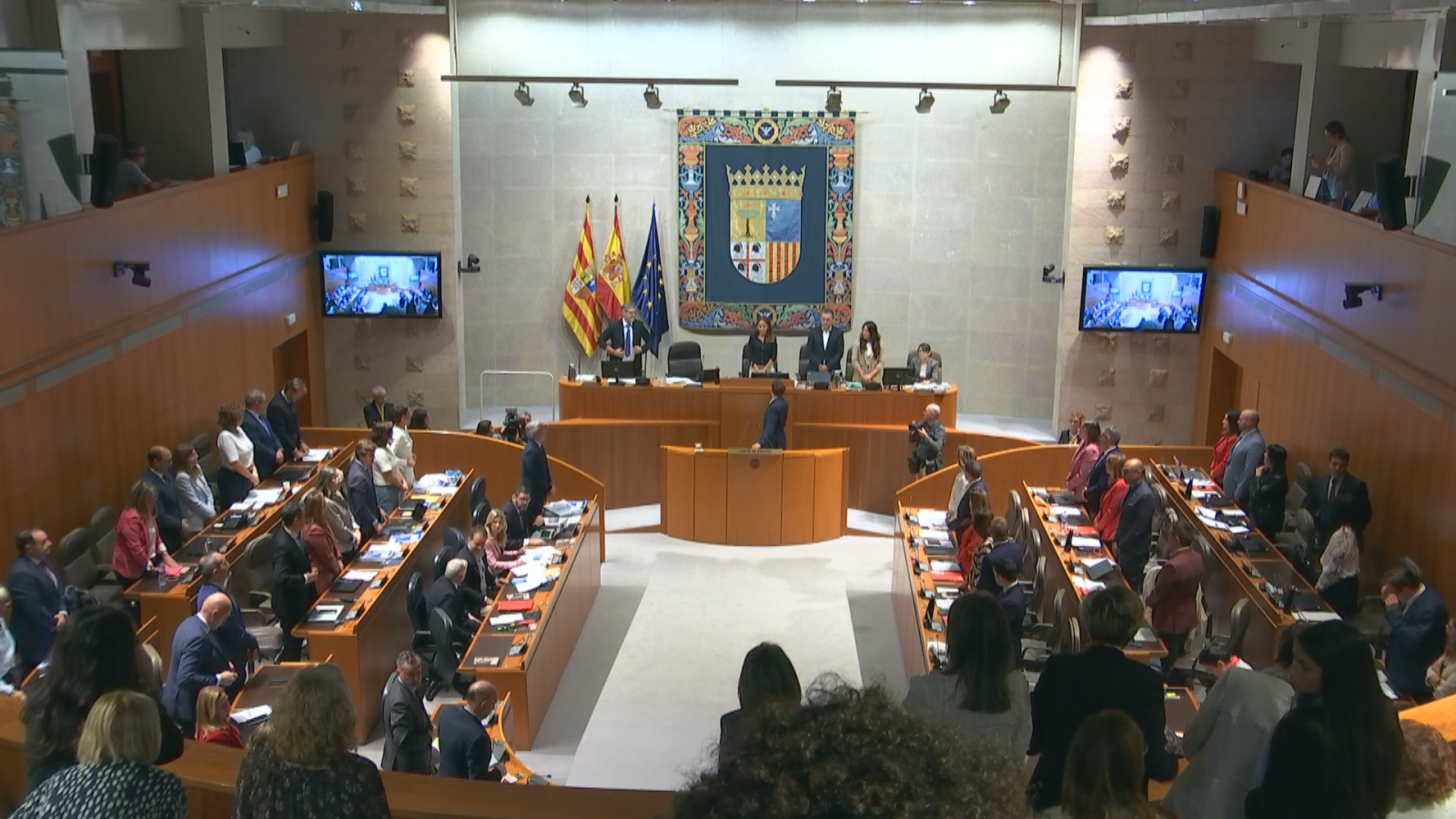 Vista general del hemiciclo de las Cortes de Aragón durante una sesión plenaria con la Mesa presidencial y los diputados en pie en las bancadas.