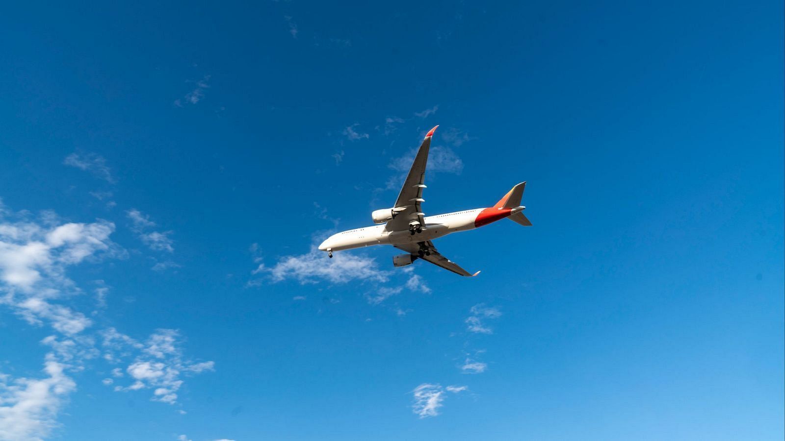 Avión comercial bimotor blanco y rojo con el tren de aterrizaje desplegado sobrevolando un cielo azul despejado.