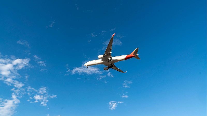 Avión comercial bimotor blanco y rojo con el tren de aterrizaje desplegado sobrevolando un cielo azul despejado.