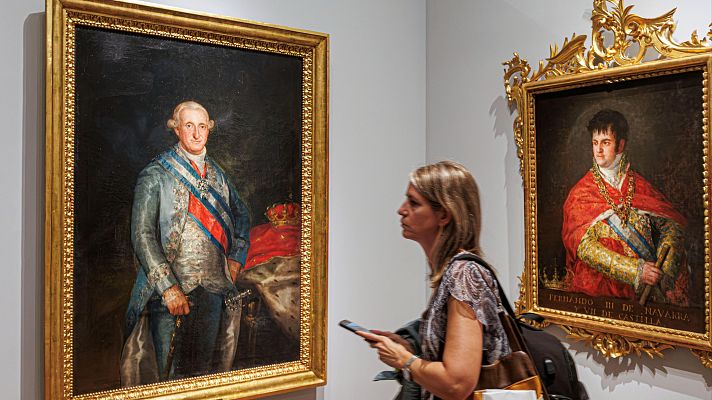Sala de museo con los retratos enmarcados en oro de Carlos IV y Fernando VII, y una visitante observando la exposición.