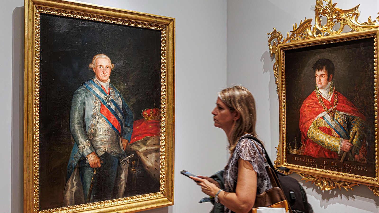 Sala de museo con los retratos enmarcados en oro de Carlos IV y Fernando VII, y una visitante observando la exposición.