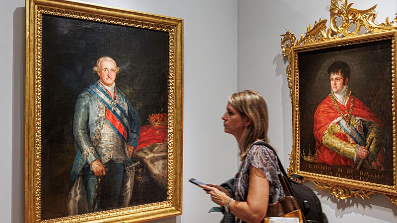 Sala de museo con los retratos enmarcados en oro de Carlos IV y Fernando VII, y una visitante observando la exposición.