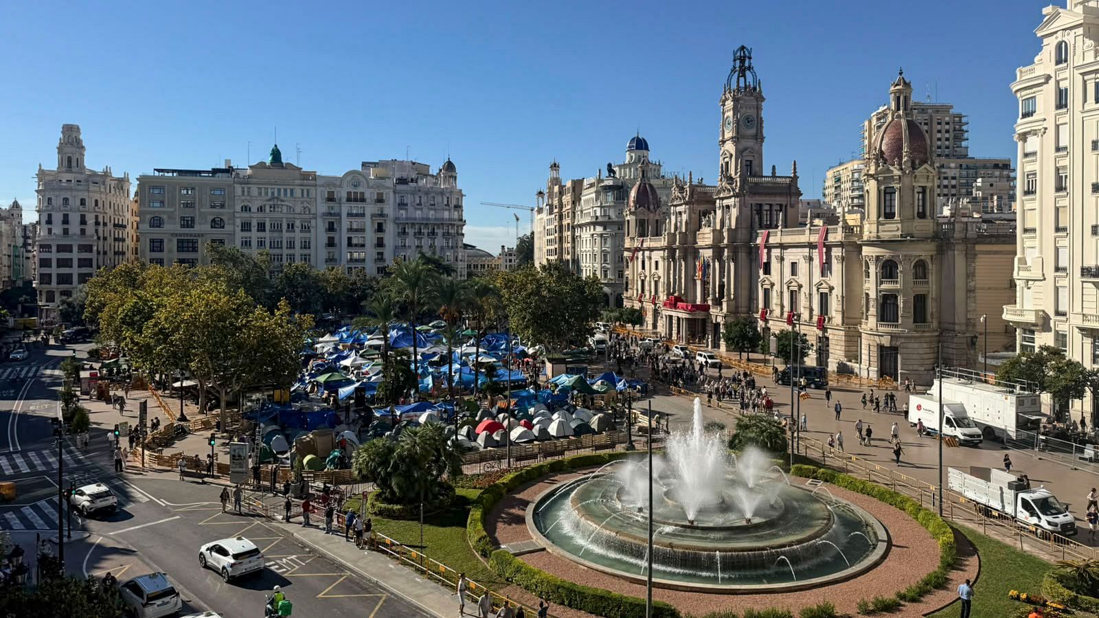 Plaza del Ayuntamiento de Valencia en un día soleado, con la Casa Consistorial, una fuente circular y un campamento de tiendas de campaña en la zona peatonal.