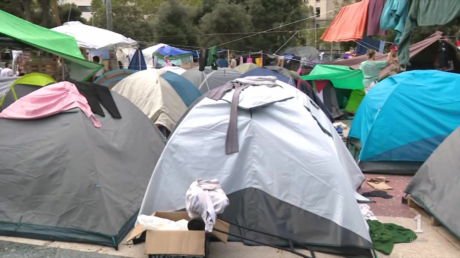 Campamento improvisado en zona urbana con tiendas de campaña, ropa tendida y personas circulando, mostrando el logotipo de La 1.