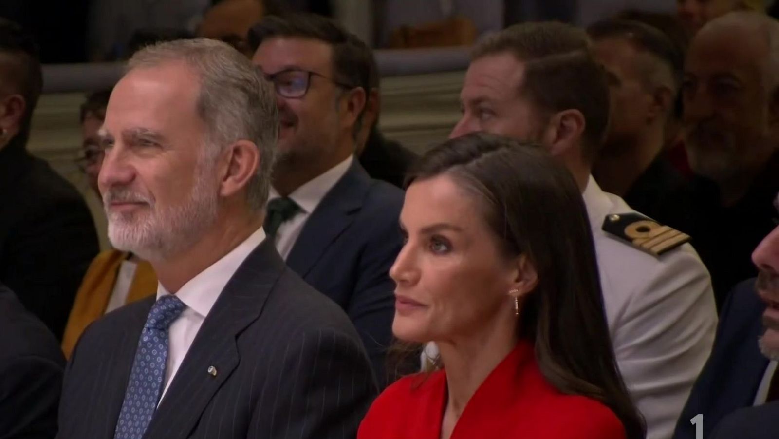 Los reyes Felipe VI y Letizia asisten atentos a un acto formal en interiores, acompañados por público y un mando militar.