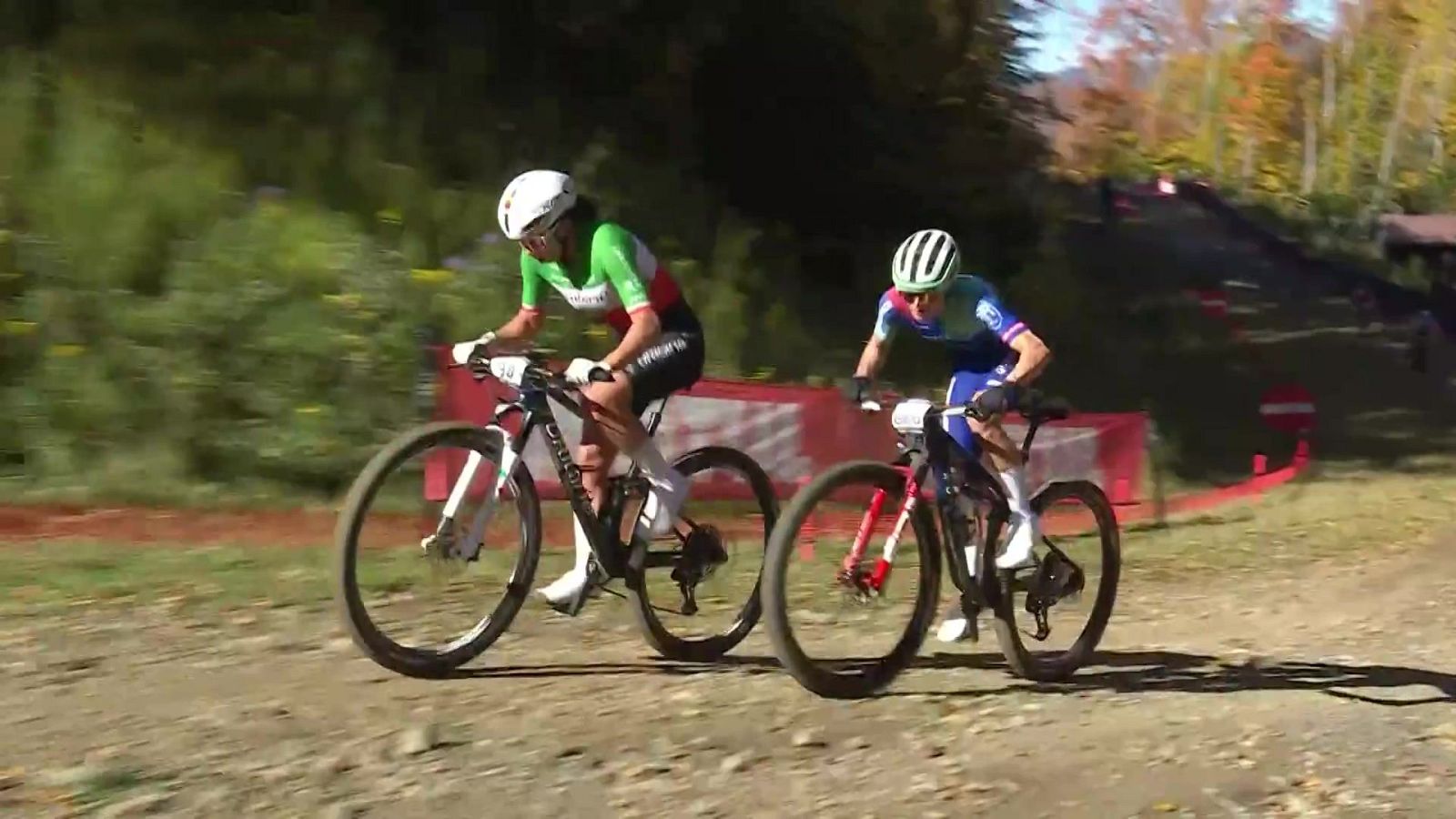 Dos ciclistas profesionales compiten en un circuito de tierra: Martina Berta con el maillot de Italia y Puck Pieterse con equipación azul, rodando concentradas sobre bicicletas de montaña en un entorno boscoso.