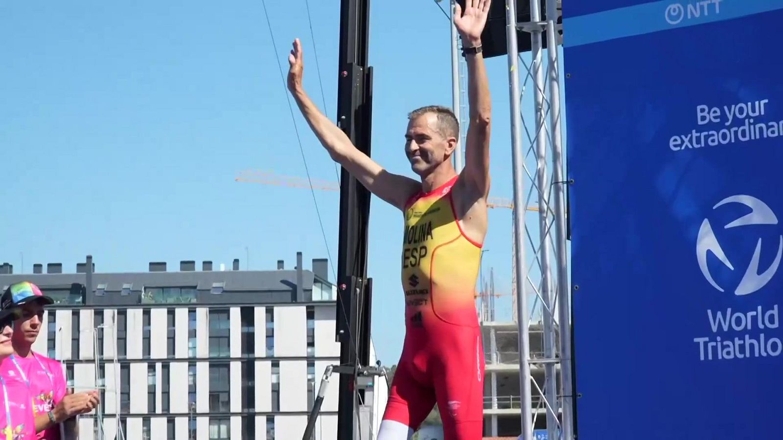 El paratriatleta Daniel Molina celebra en el podio con los brazos en alto, vistiendo el mono de la selección española de triatlón bajo un cielo azul.