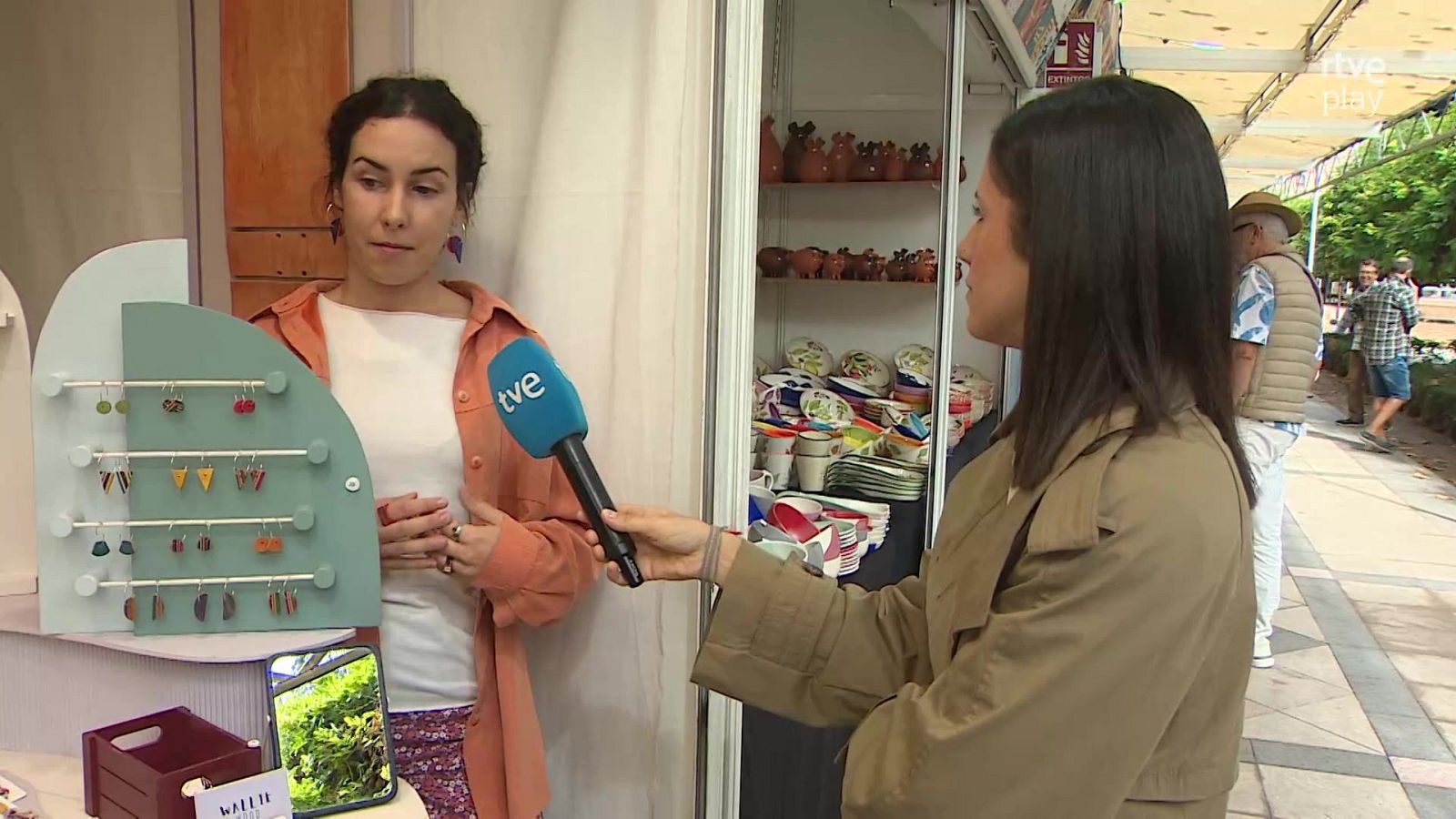 Una reportera con gabardina caqui entrevista a una artesana en una feria al aire libre con expositores de joyería y cerámica.