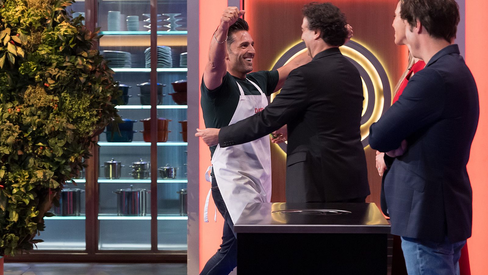 Un concursante celebra su triunfo levantando el puño mientras se abraza con un juez en el plató de un programa de cocina.