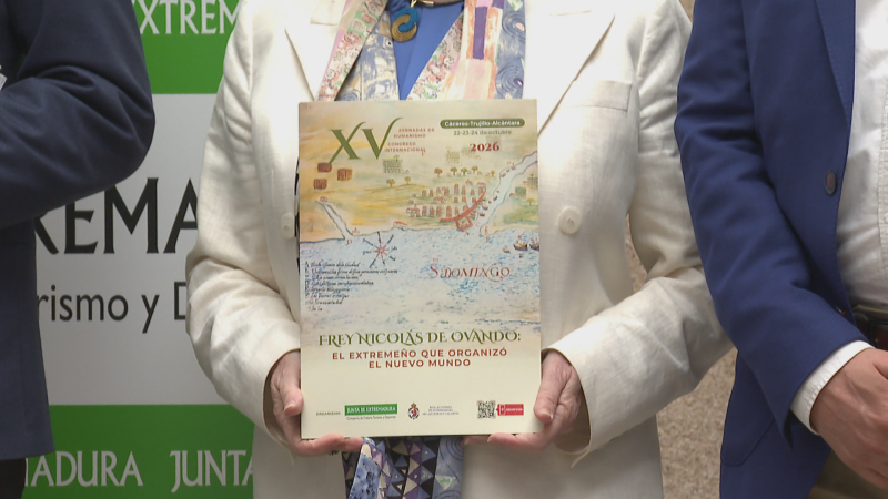 Mujer sosteniendo el cartel de un congreso cultural junto a dos personas, con un expositor de Extremadura al fondo.