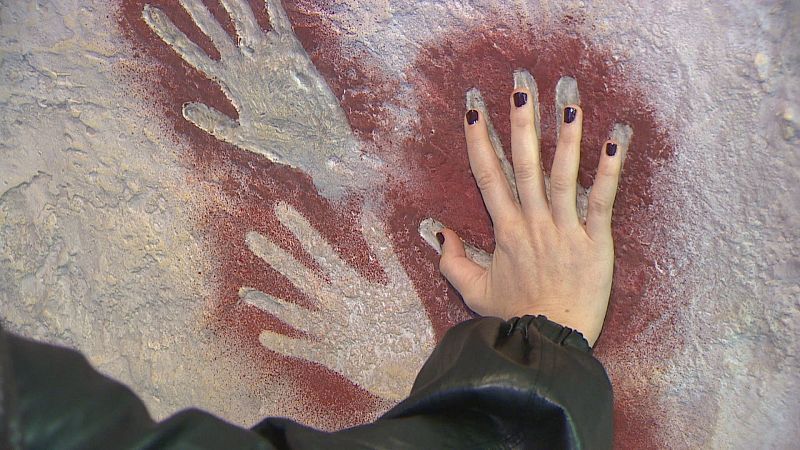 Mano interactuando con una réplica táctil de arte rupestre con siluetas de manos en negativo hechas con pigmento rojo.