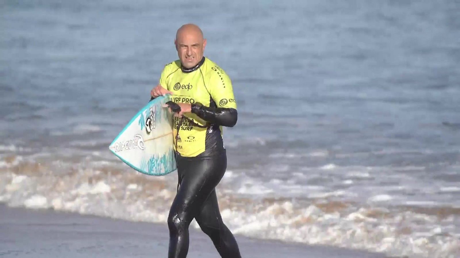 Cristóbal Soria camina por la playa con neopreno negro, licra amarilla y una tabla de surf bajo el brazo.