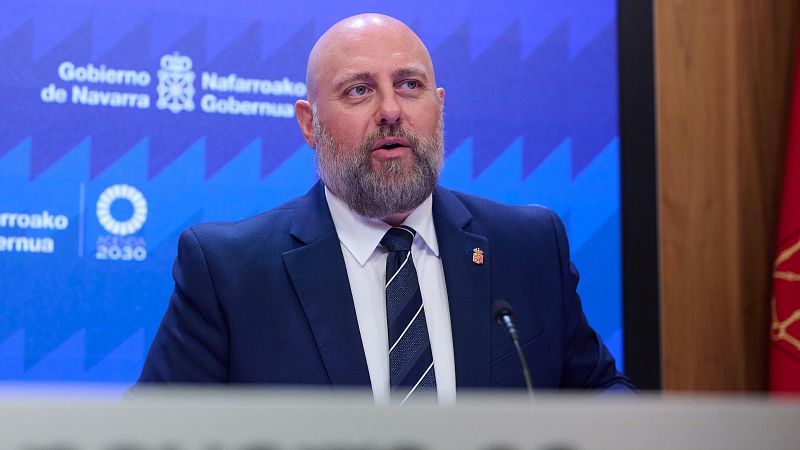Un hombre con traje y corbata comparece en una rueda de prensa ante un atril, con fondo azul institucional y la bandera de Navarra a un lado.