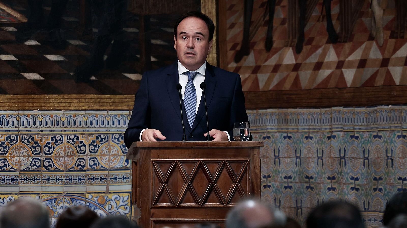 Hombre de traje azul y corbata clara interviene ante un atril de madera con dos micrófonos en una sala con zócalos de azulejos tradicionales y un cuadro al fondo.