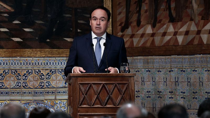 Hombre de traje azul y corbata clara interviene ante un atril de madera con dos micrófonos en una sala con zócalos de azulejos tradicionales y un cuadro al fondo.