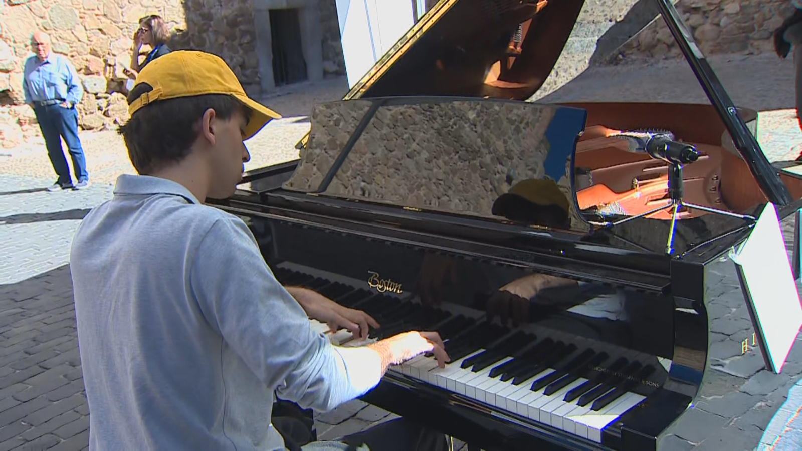 Joven con gorra amarilla tocando un piano de cola negro al aire libre en una plaza adoquinada bajo la luz del sol.