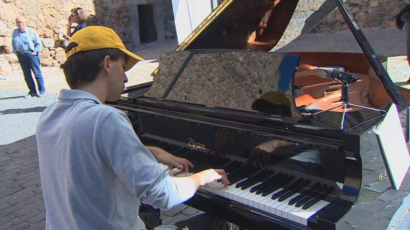 Joven con gorra amarilla tocando un piano de cola negro al aire libre en una plaza adoquinada bajo la luz del sol.
