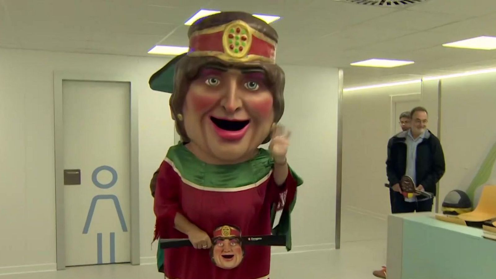 La Pilara, el tradicional cabezudo zaragozano, posa alegremente en un pasillo interior con vestimenta típica y una barra en las manos.