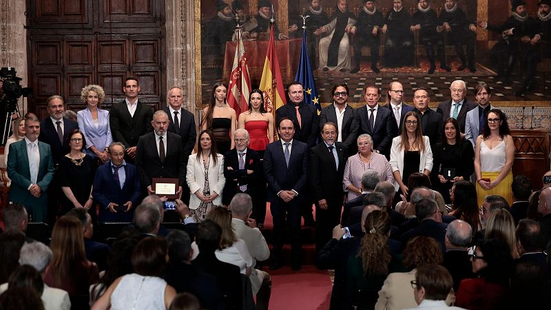 Foto de familia de la entrega de Altas Distinciones de la Generalitat con Carlos Mazón y los galardonados ante un mural histórico y banderas oficiales.