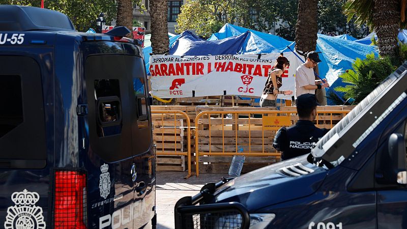 Furgones policiales y un agente frente a una acampada de protesta delimitada por vallas y una pancarta política en una plaza.