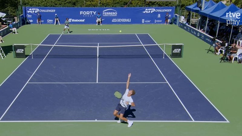 Plano general de un partido de tenis al aire libre donde un jugador se prepara para sacar en una pista de suelo azul y verde con jueces y espectadores al fondo.