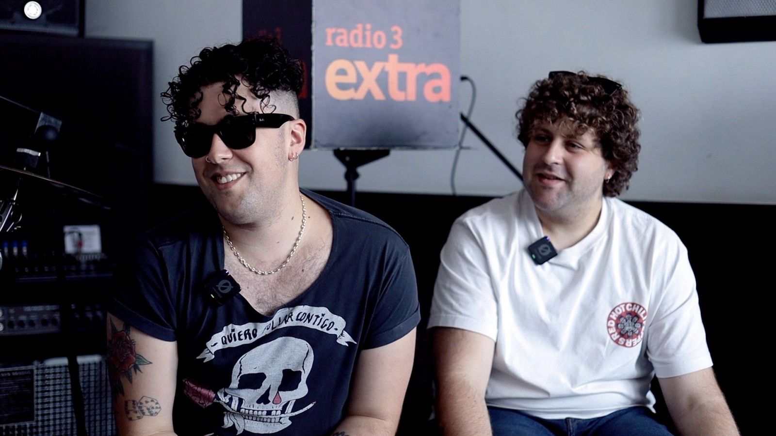 Dos j�venes conversan sonrientes en un estudio de grabaci�n, uno con gafas de sol y camiseta estampada y el otro con camiseta blanca y micr�fono de solapa.