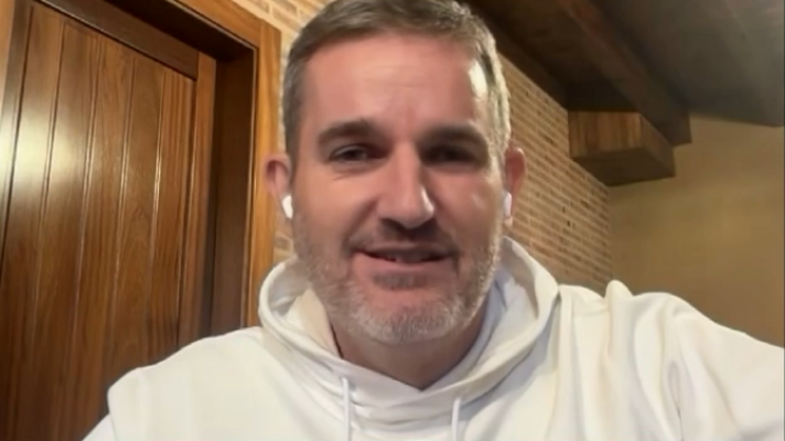 Hombre de mediana edad con barba canosa y sudadera blanca con capucha, en una videollamada frente a una pared de ladrillo visto.