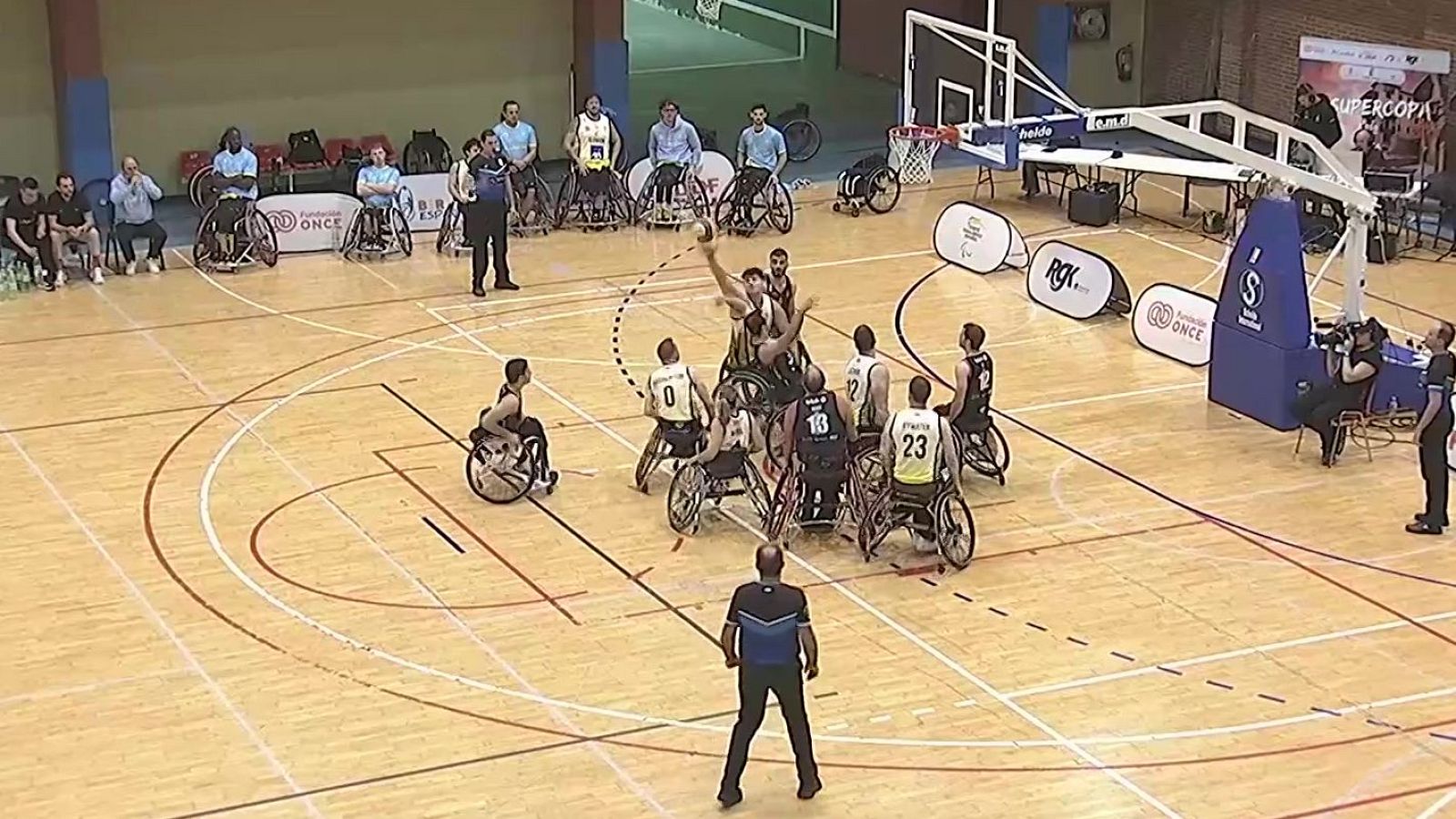 Intensa jugada de un partido de baloncesto en silla de ruedas en un pabellón con pista de parqué y vallas publicitarias.