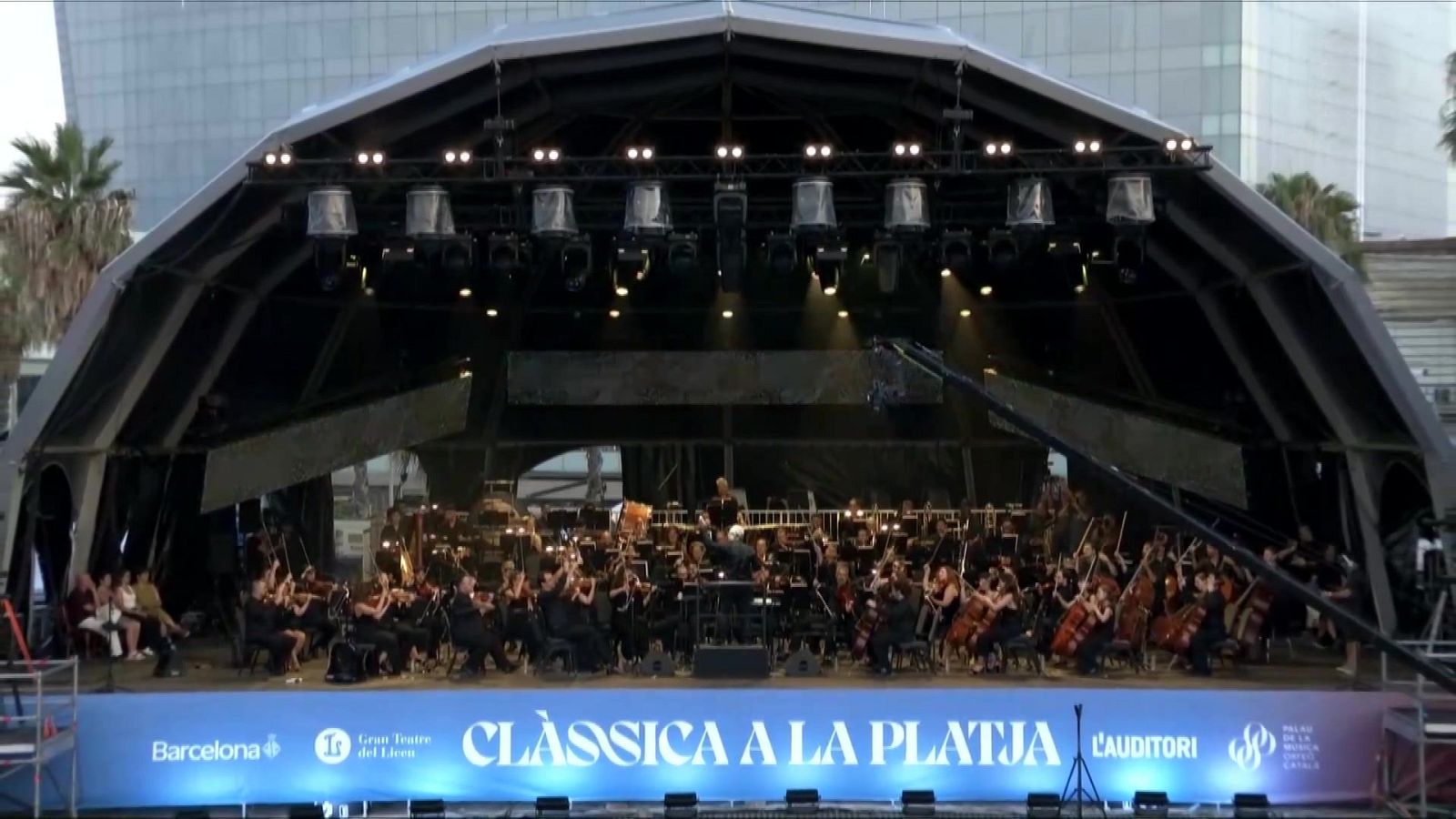 Orquesta sinfónica interpretando un concierto al aire libre en un gran escenario iluminado bajo una estructura abovedada.