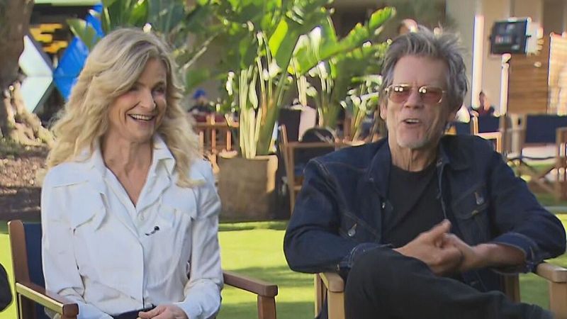 Los actores Kyra Sedgwick y Kevin Bacon sentados al aire libre en sillas de madera sobre césped verde durante una entrevista.