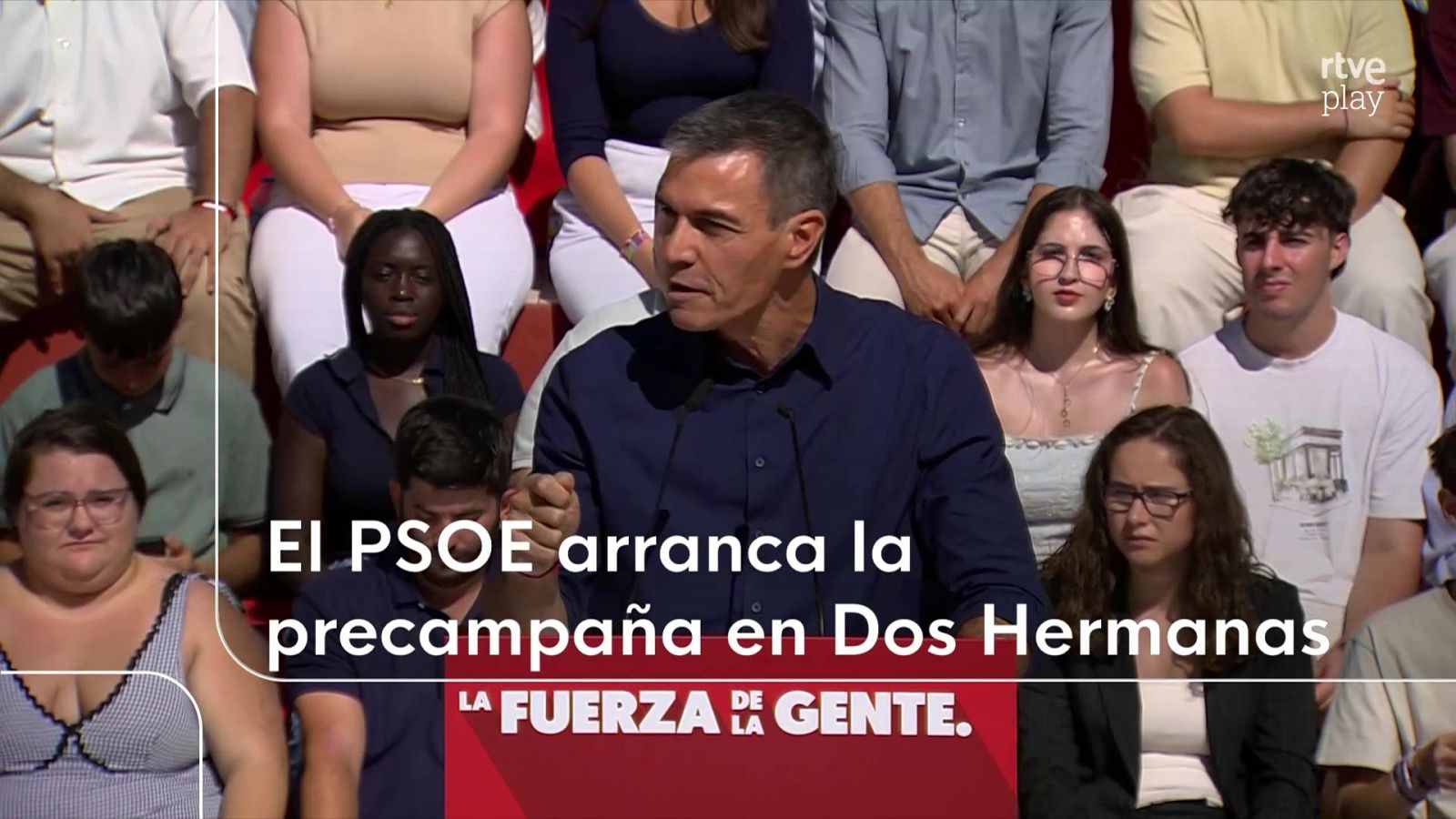 Pedro Sánchez interviene con expresión seria ante dos micrófonos en un acto público retransmitido por televisión.