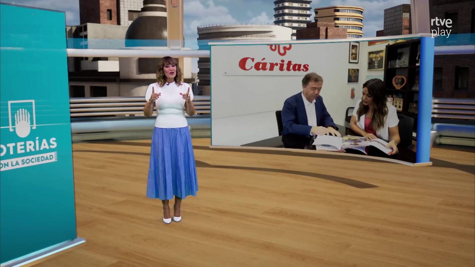 La presentadora Sandra Daviú en el plató del programa, vistiendo blusa blanca y falda azul, sonriendo ante una pantalla con imágenes de Cáritas y un fondo urbano.