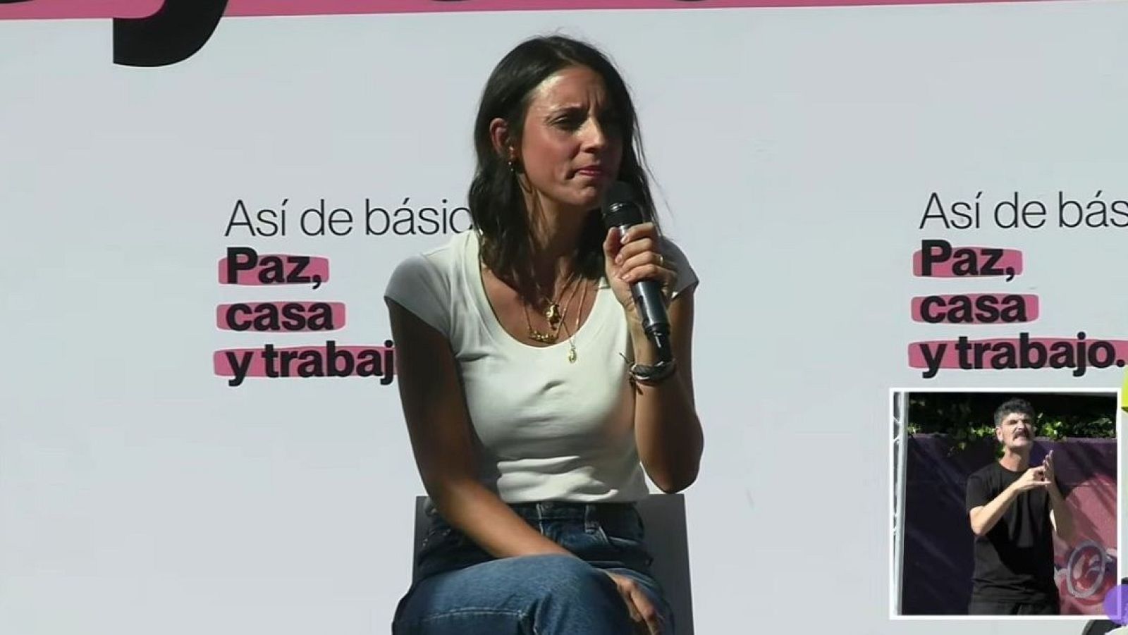 Irene Montero interviene en un acto público sosteniendo un micrófono, con un panel de fondo y un intérprete de lengua de signos en la esquina.