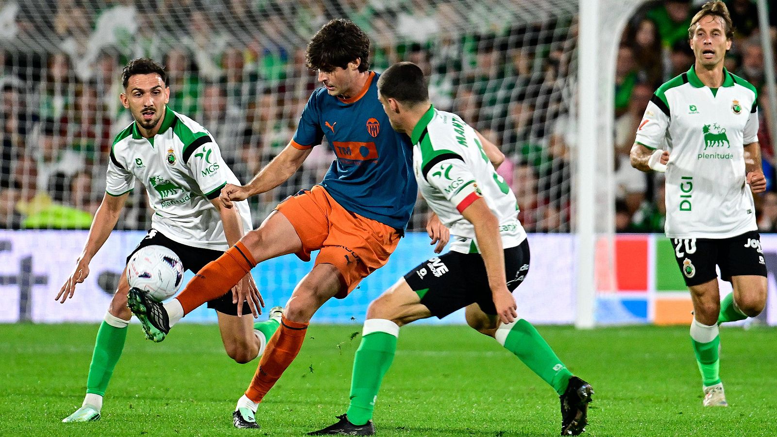 Futbolista con camiseta azul marino y naranja intenta rematar un balón a media altura, presionado por varios defensas rivales de blanco y negro cerca de la portería.