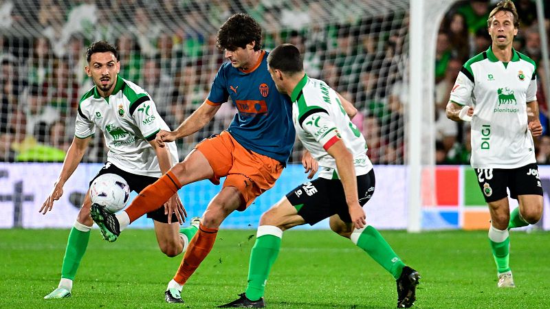 Futbolista con camiseta azul marino y naranja intenta rematar un balón a media altura, presionado por varios defensas rivales de blanco y negro cerca de la portería.