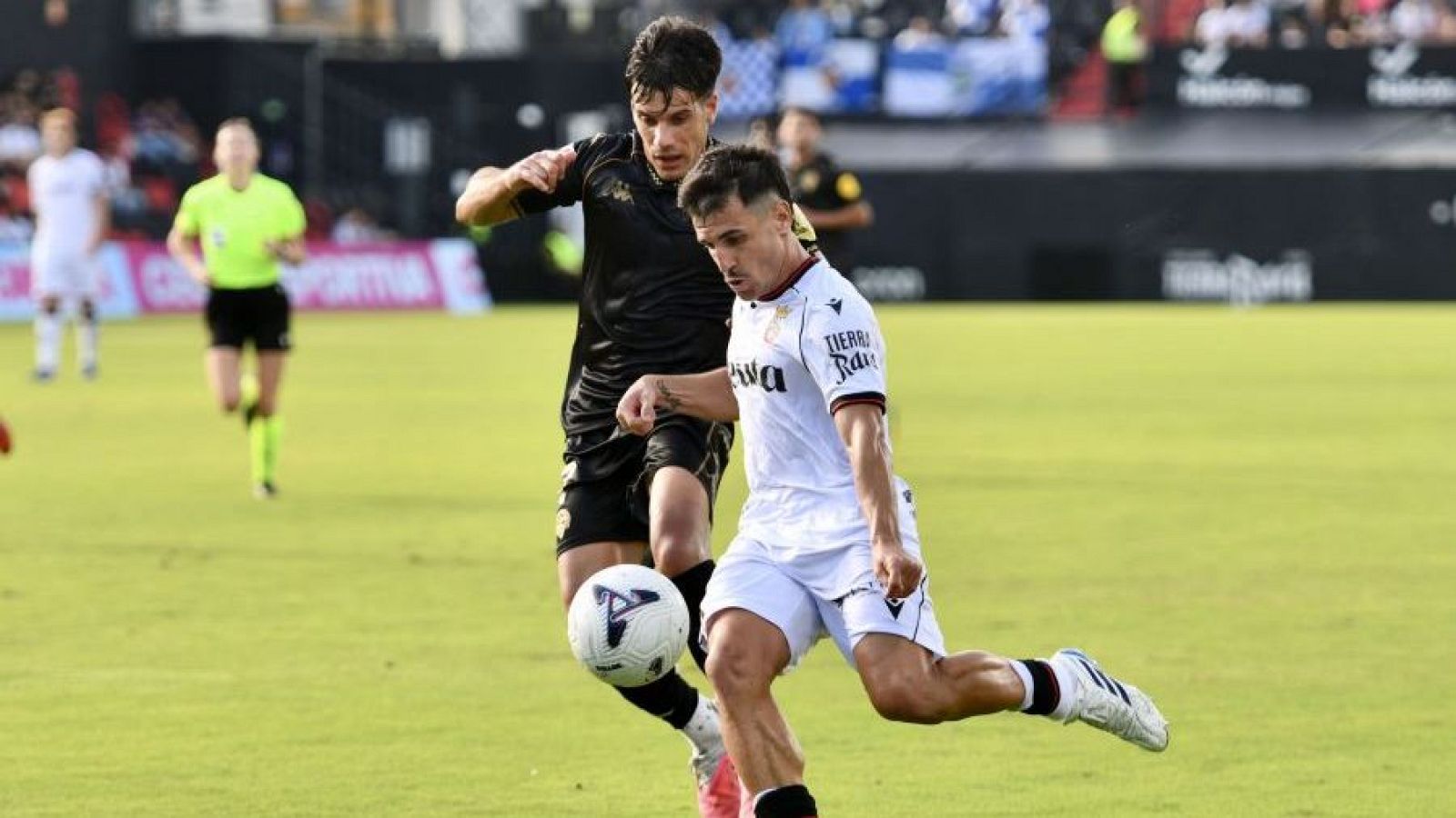 Intensa disputa por el balón entre un futbolista de blanco y un rival de negro durante un partido de fútbol en un estadio.
