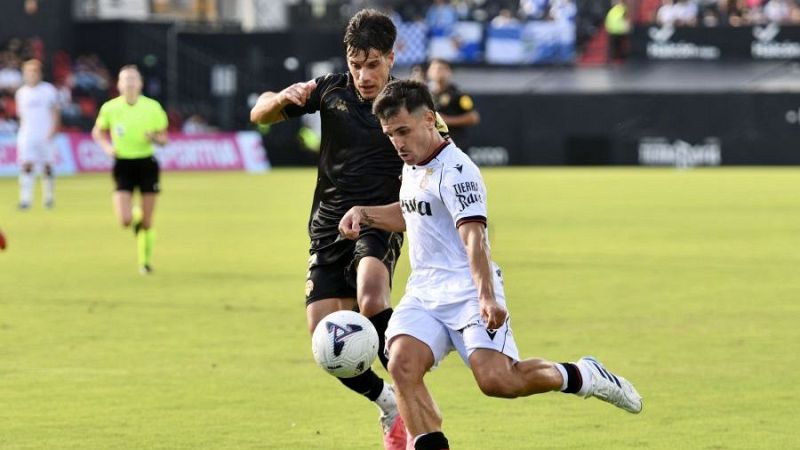 Intensa disputa por el bal�n entre un futbolista de blanco y un rival de negro durante un partido de f�tbol en un estadio.