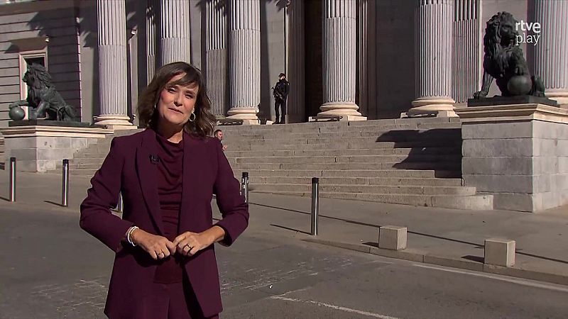 Una presentadora con traje burdeos y micrófono de solapa se encuentra frente al Congreso de los Diputados en Madrid, con el logotipo de RTVE Play visible.