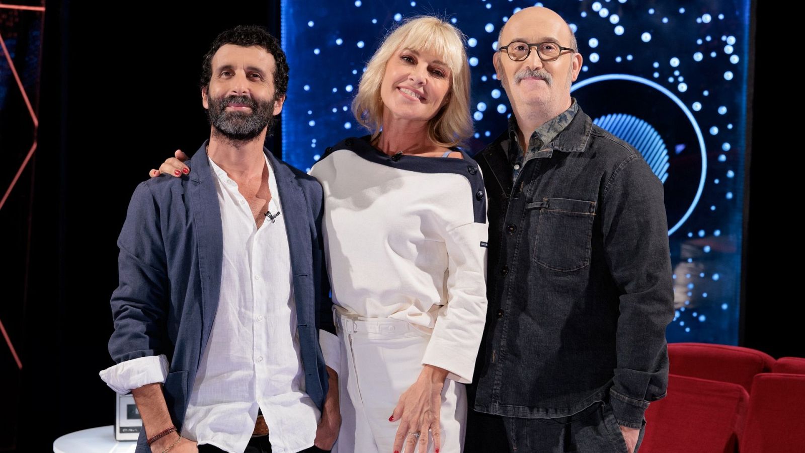 Tres conocidos profesionales del cine y la televisión posan sonrientes y cercanos en un plató con iluminación azul y butacas rojas.