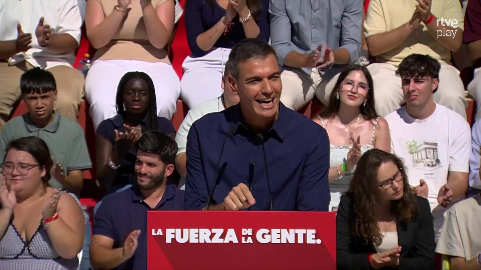 Pedro Sánchez habla sonriente ante micrófonos junto a un atril rojo con la frase «LA FUERZA DE LA GENTE.» en un acto político con público al fondo.