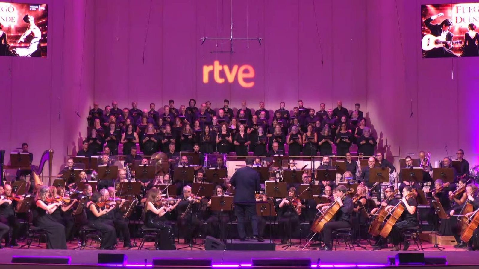 Actuación en directo de una orquesta sinfónica y coro en un escenario iluminado en tonos magenta y violeta con el logotipo de RTVE al fondo.