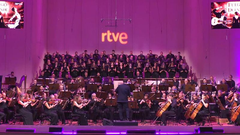 Actuación en directo de una orquesta sinfónica y coro en un escenario iluminado en tonos magenta y violeta con el logotipo de RTVE al fondo.