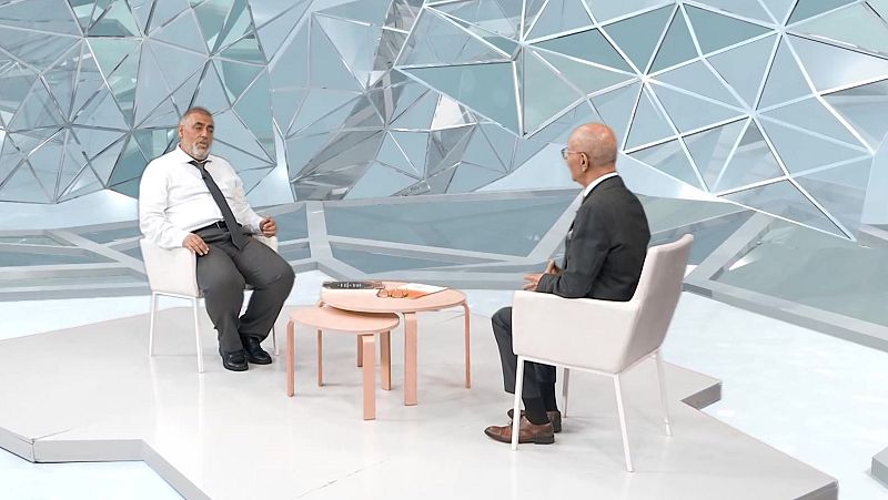 Plató de televisión moderno con paneles geométricos donde un presentador y un invitado conversan sentados en sillones blancos.