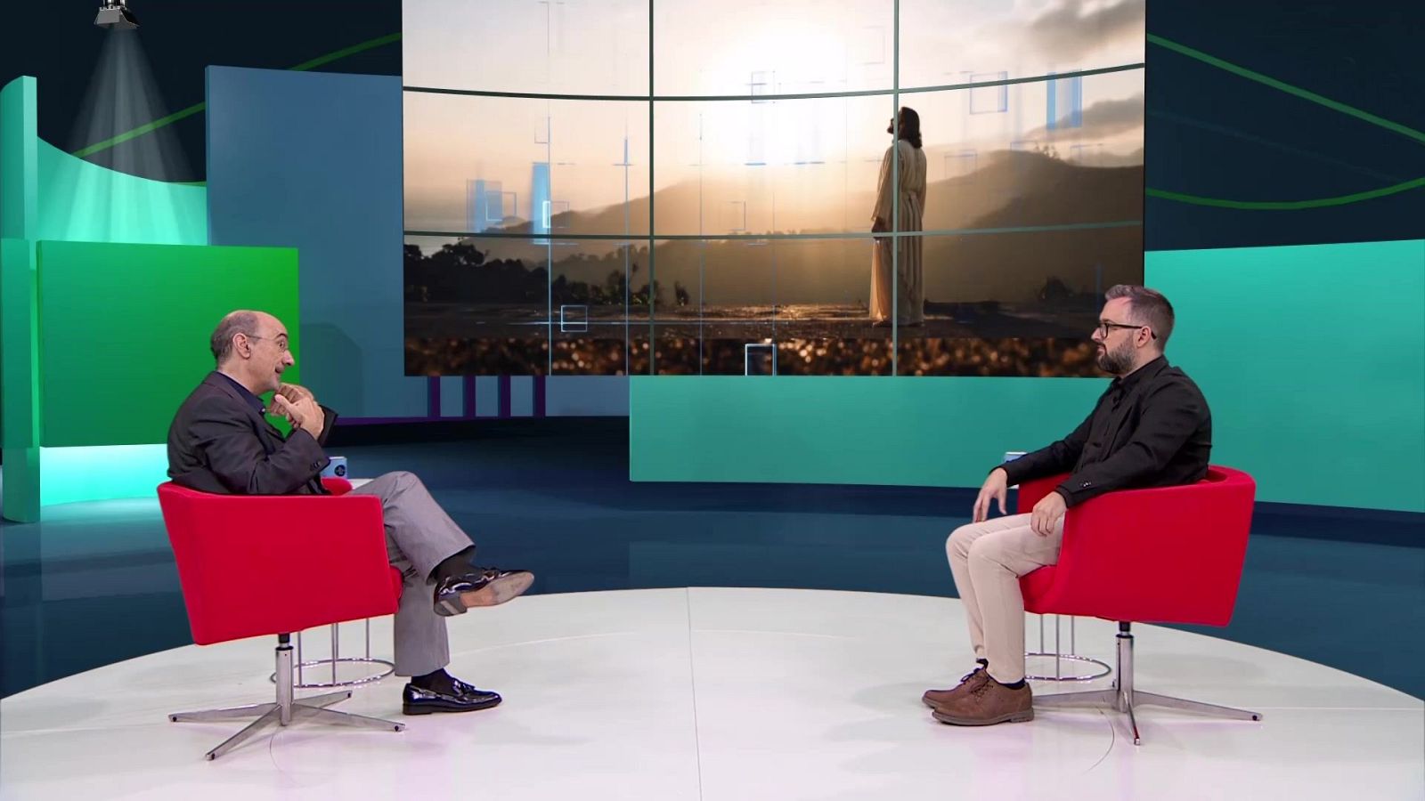 Dos hombres sentados en sillones rojos dialogan en un plató de televisión con paneles verdes y azules y un videowall de fondo.