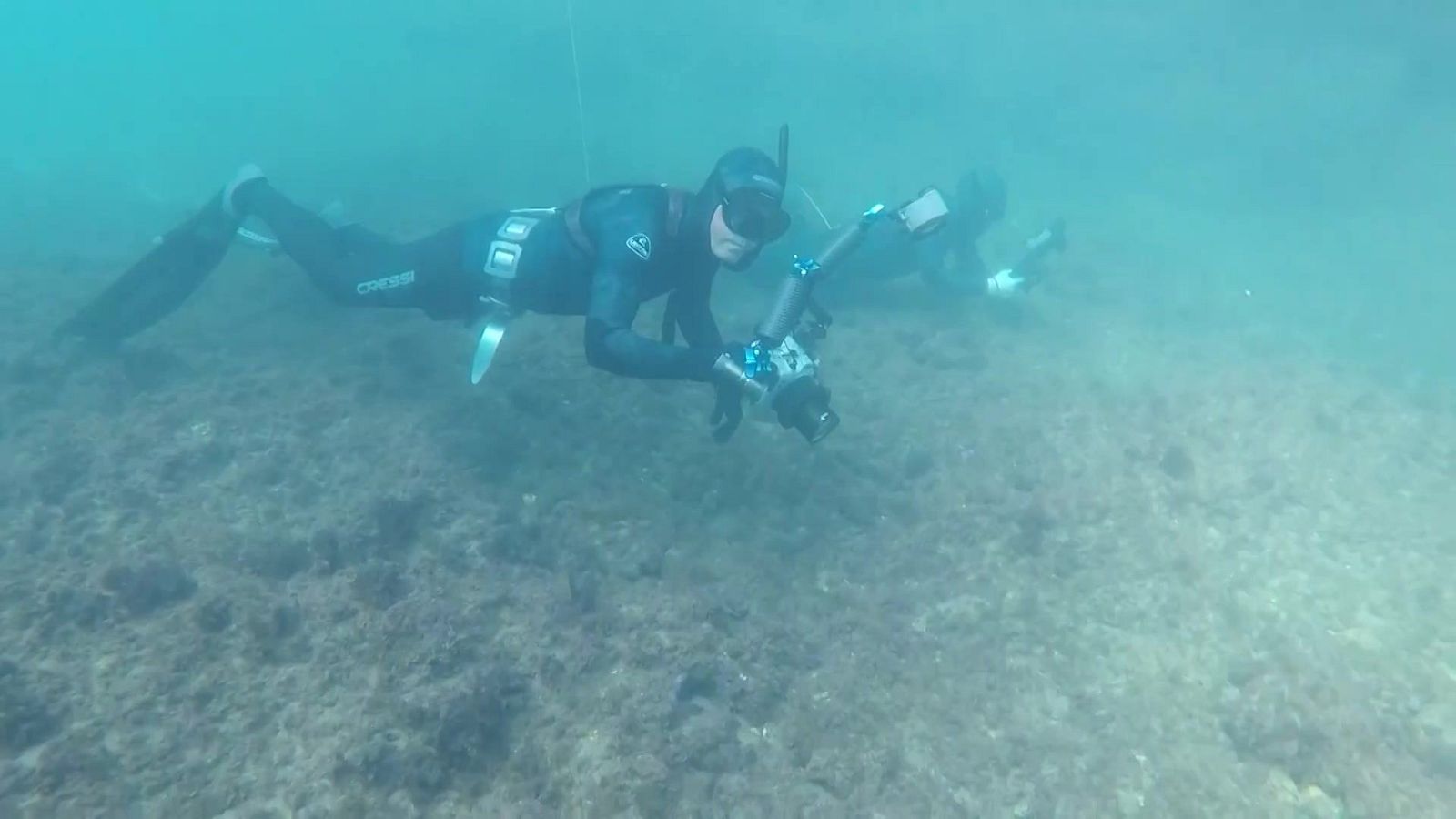Dos submarinistas con neopreno negro y cámaras acuáticas realizan fotografía submarina en apnea sobre un lecho rocoso y arenoso.