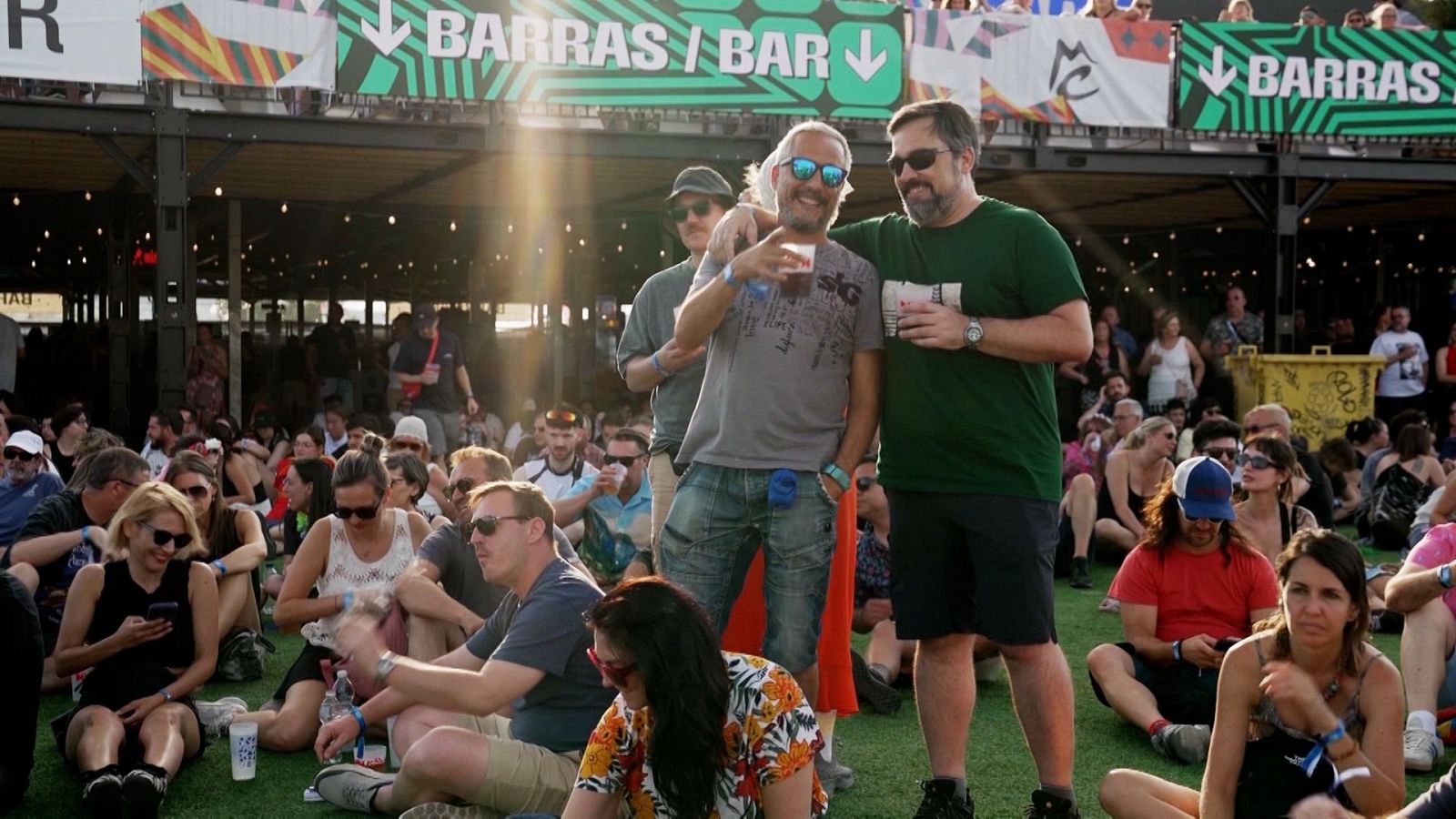 Dos hombres sonrientes abrazados con vasos de plástico en un festival al aire libre con césped artificial y asistentes descansando.