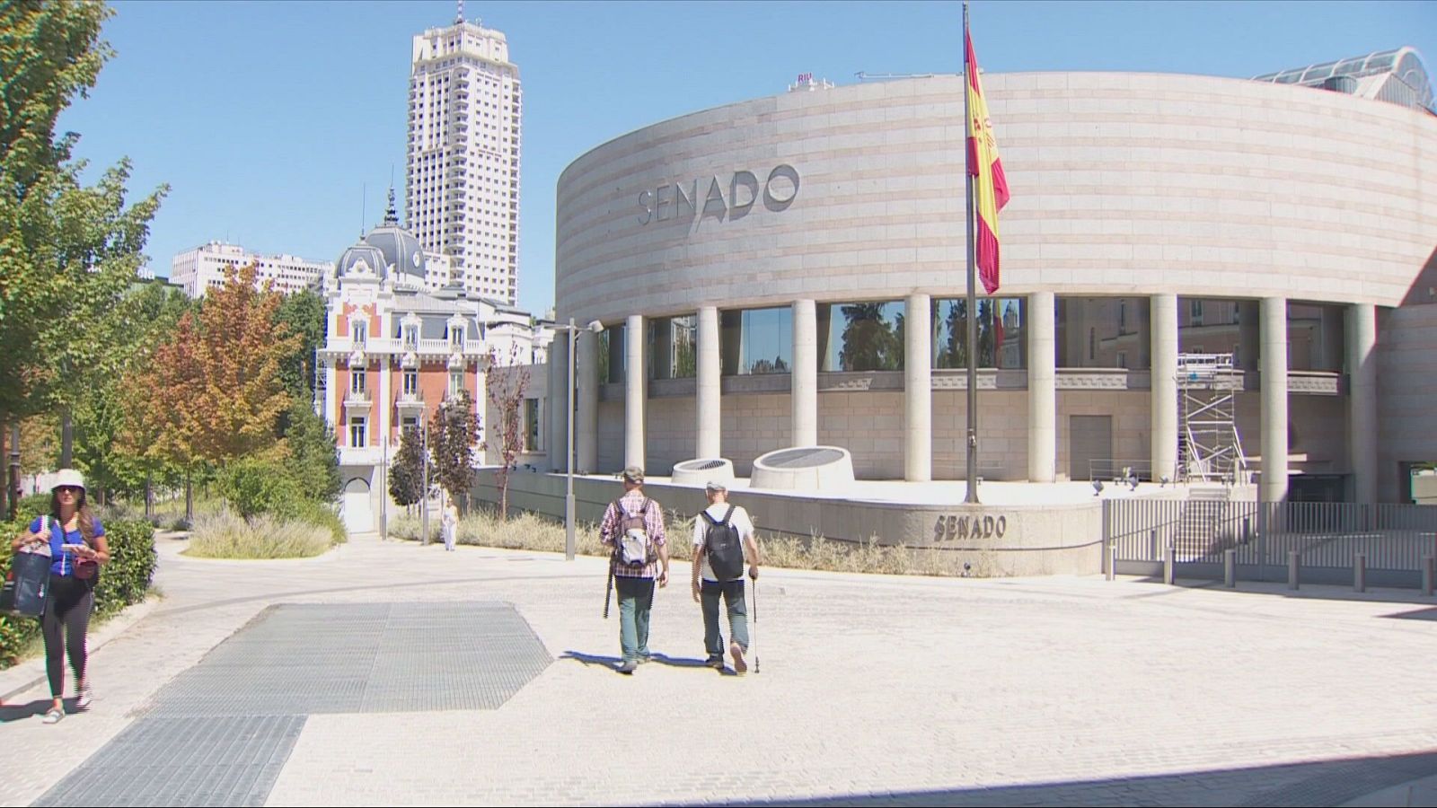 Fachada curva del edificio del Senado de España en Madrid en un día soleado, con una plaza peatonal en primer plano y la bandera nacional ondeando en un mástil.